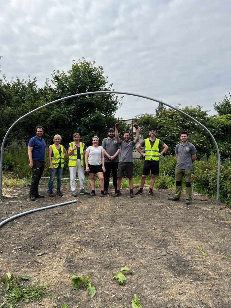 A group of happy people dressed in gardening wear stood in a line outside under a polytunnel arch.