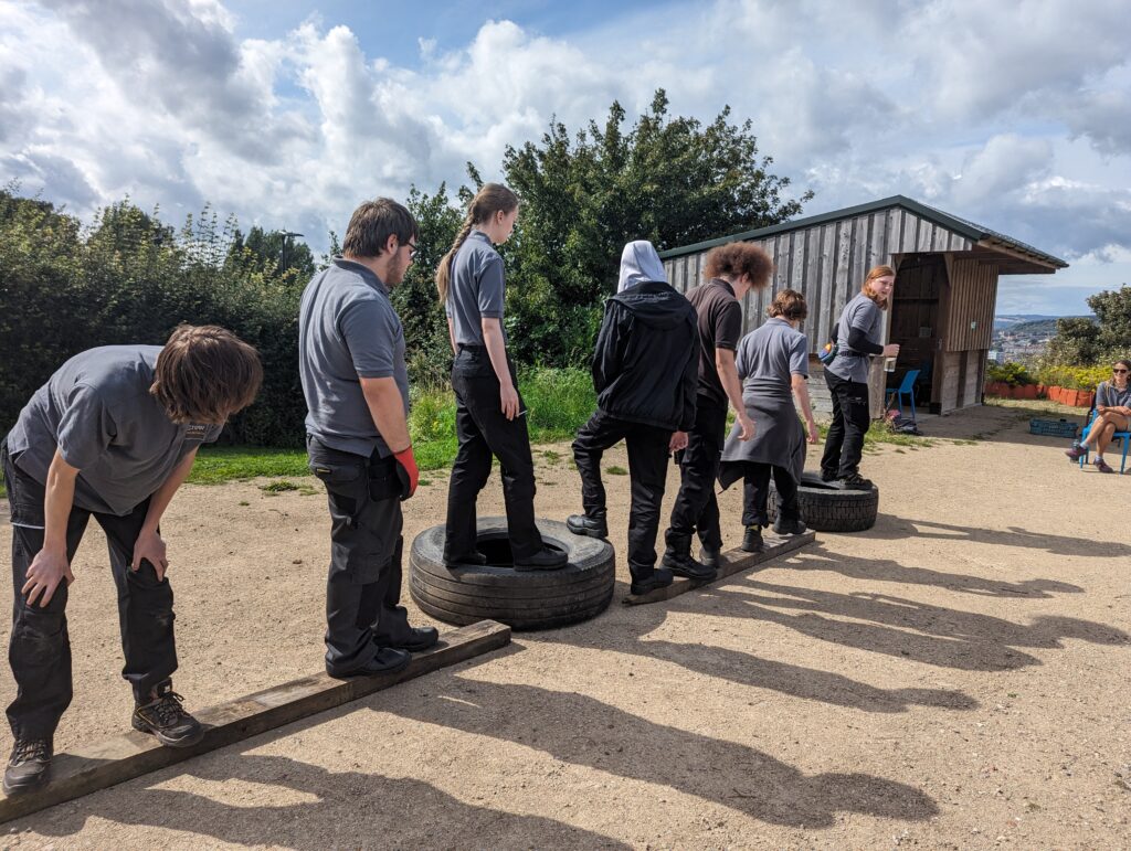 A group of people team building along an obstacle of wooden planks and tyres.