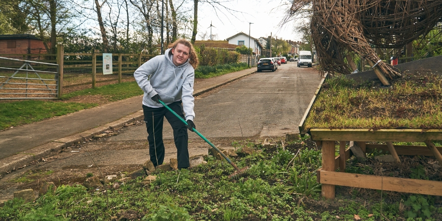 Young male using a gardening rake outdoors with a road in the background.