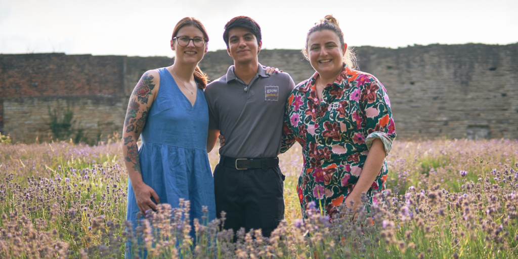 Two females and a male stood smiling, arm-in-arm, in a meadow field.